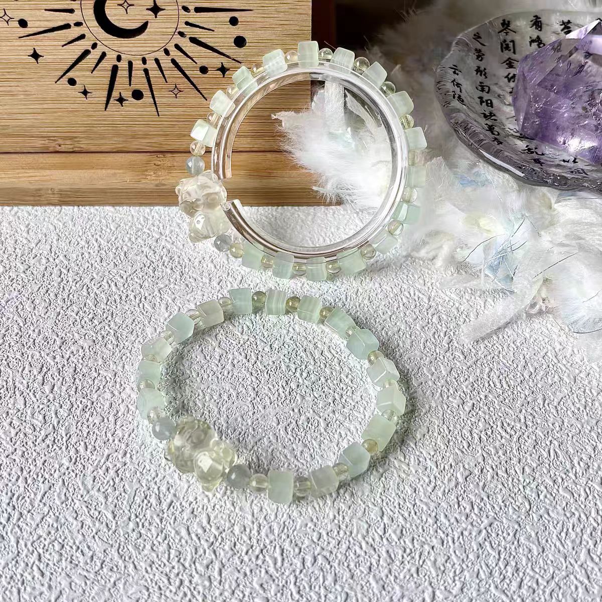 Two beaded bracelets on a textured surface with a wooden board and decorative items in the background.