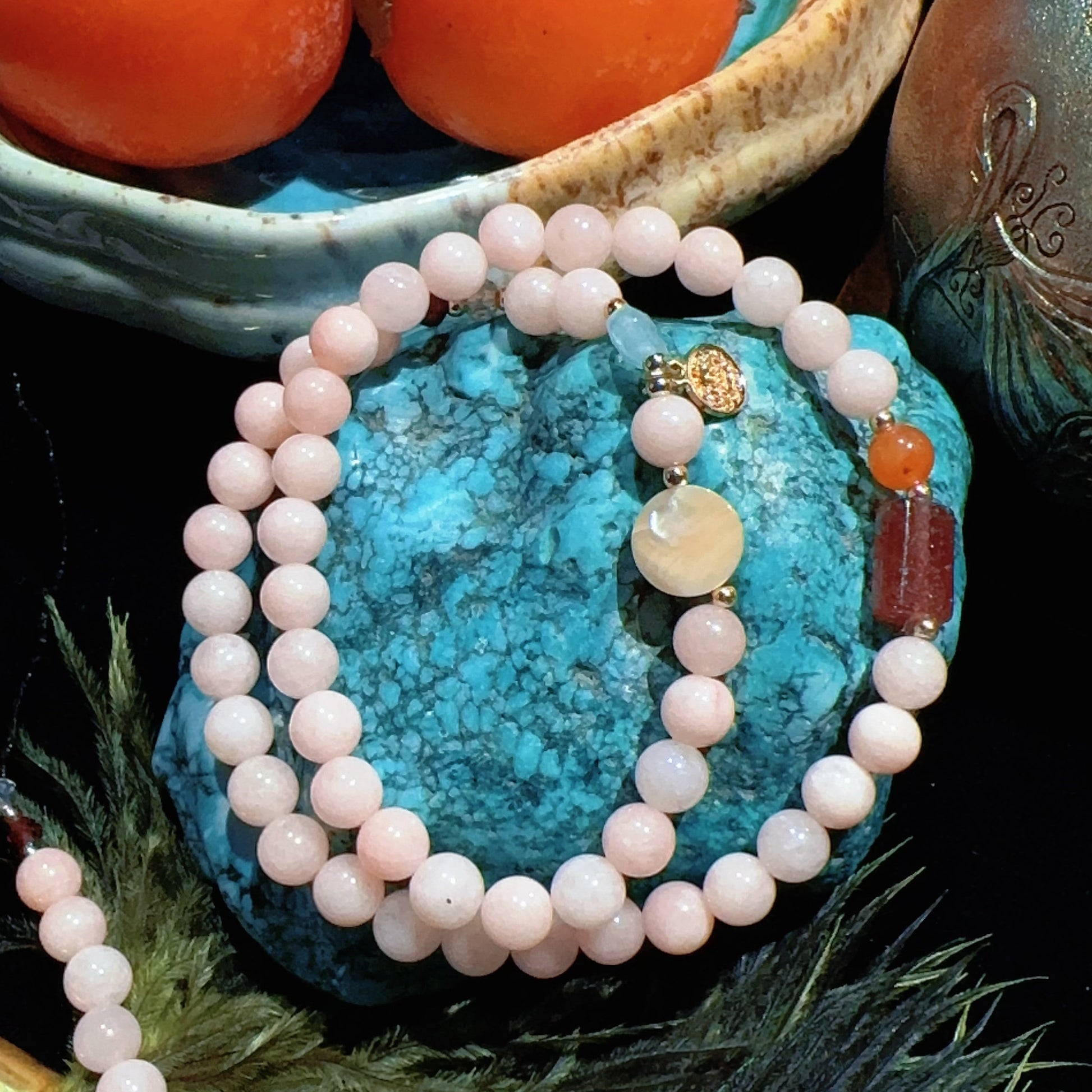 Necklace with white beads on a turquoise stone, surrounded by fruits and a teapot.